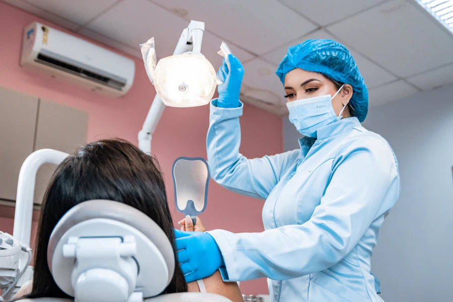 Una dentista con gorro y guantes azules sostiene un espejo de mano para mostrarle los dientes a una paciente sentada.